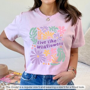 K&ouml;nnte beinhalten: Hellrosa T-Shirt mit dem Text "Live Like Wildflowers" in Gr&uuml;n und Lila, umgeben von bunten Blumenillustrationen. Das T-Shirt hat eine hellrosa Farbe.