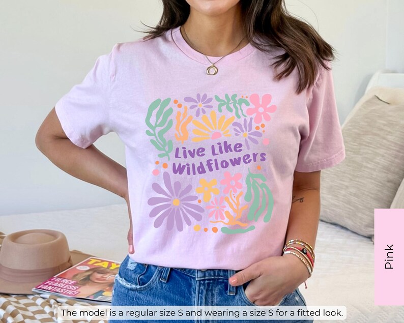K&ouml;nnte beinhalten: Hellrosa T-Shirt mit dem Text "Live Like Wildflowers" in Gr&uuml;n und Lila, umgeben von bunten Blumenillustrationen. Das T-Shirt hat eine hellrosa Farbe.