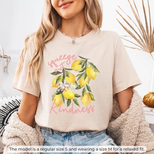 K&ouml;nnte beinhalten: Ein beiges T-Shirt mit einem Motiv aus Zitronen und Bl&auml;ttern. Der Text "squeeze out kindness" ist in Rosa auf das Shirt gedruckt.