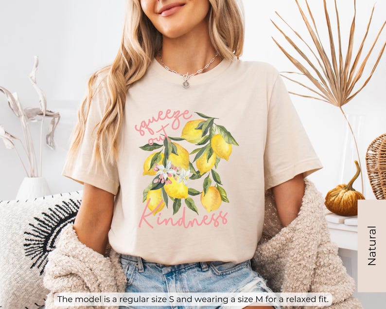 K&ouml;nnte beinhalten: Ein beiges T-Shirt mit einem Motiv aus Zitronen und Bl&auml;ttern. Der Text "squeeze out kindness" ist in Rosa auf das Shirt gedruckt.