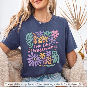 K&ouml;nnte beinhalten: Marineblaues T-Shirt mit floralem Design und dem Text "Live Like Wildflowers" in einer farbenfrohen, verspielten Schriftart. Das Shirt ist in der Gr&ouml;&szlig;e S erh&auml;ltlich, und das Model tr&auml;gt Gr&ouml;&szlig;e M f&uuml;r eine lockere Passform.