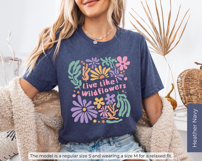 K&ouml;nnte beinhalten: Marineblaues T-Shirt mit floralem Design und dem Text "Live Like Wildflowers" in einer farbenfrohen, verspielten Schriftart. Das Shirt ist in der Gr&ouml;&szlig;e S erh&auml;ltlich, und das Model tr&auml;gt Gr&ouml;&szlig;e M f&uuml;r eine lockere Passform.