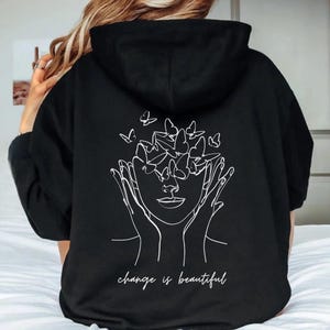 May include: Black hooded sweatshirt with a white graphic of a face with butterflies around it. The text "change is beautiful" is printed below the graphic.
