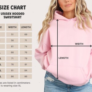 May include: Size chart for a unisex hooded sweatshirt. The chart shows the width and length measurements in centimeters for sizes S through 5XL. A model is wearing a pink hooded sweatshirt in size XL.
