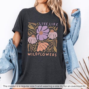May include: Dark grey heather t-shirt with a floral design and the text "LIVE LIKE WILDFLOWERS" in a retro font. The design features purple, peach, and white flowers with brown leaves. The model is wearing a size XL for an oversized fit.
