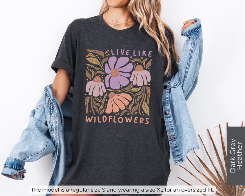 May include: Dark grey heather t-shirt with a floral design and the text "LIVE LIKE WILDFLOWERS" in a retro font. The design features purple, peach, and white flowers with brown leaves. The model is wearing a size XL for an oversized fit.