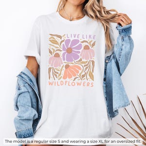 May include: White t-shirt with a floral design in shades of purple, peach, and tan, with the text "LIVE LIKE WILDFLOWERS". The shirt has short sleeves and a relaxed fit, perfect for casual wear.