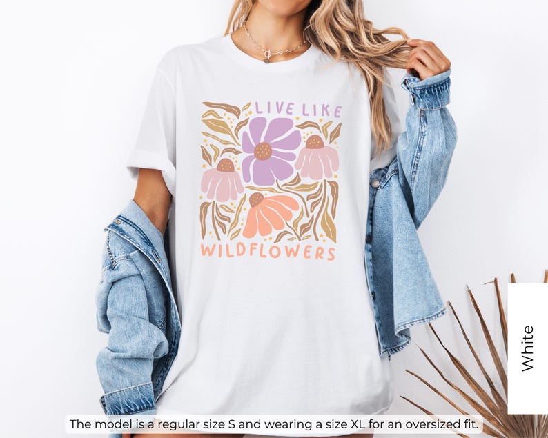 May include: White t-shirt with a floral design in shades of purple, peach, and tan, with the text "LIVE LIKE WILDFLOWERS". The shirt has short sleeves and a relaxed fit, perfect for casual wear.