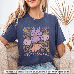 May include: Heather navy blue t-shirt with a floral design and the text "LIVE LIKE WILDFLOWERS" in a whimsical font. The design features purple, peach, and cream-colored flowers with green and gold leaves. The model is wearing a size M for a relaxed fit.
