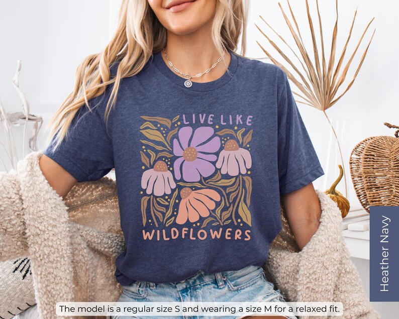 May include: Heather navy blue t-shirt with a floral design and the text "LIVE LIKE WILDFLOWERS" in a whimsical font. The design features purple, peach, and cream-colored flowers with green and gold leaves. The model is wearing a size M for a relaxed fit.