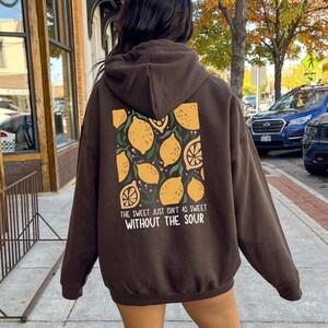 K&ouml;nnte beinhalten: Ein brauner Hoodie mit einem Zitronendruck und dem Text "The sweet just isn't as sweet without the sour" auf der R&uuml;ckseite. Dunkle Schokolade.