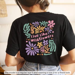 K&ouml;nnte beinhalten: Schwarzes T-Shirt mit einem bunten Blumendesign auf dem R&uuml;cken, mit dem Text "Live Like Wildflowers". Das Design umfasst Blumen in den Farbt&ouml;nen Rosa, Lila, Orange und Gr&uuml;n. Das Model tr&auml;gt Gr&ouml;&szlig;e S.