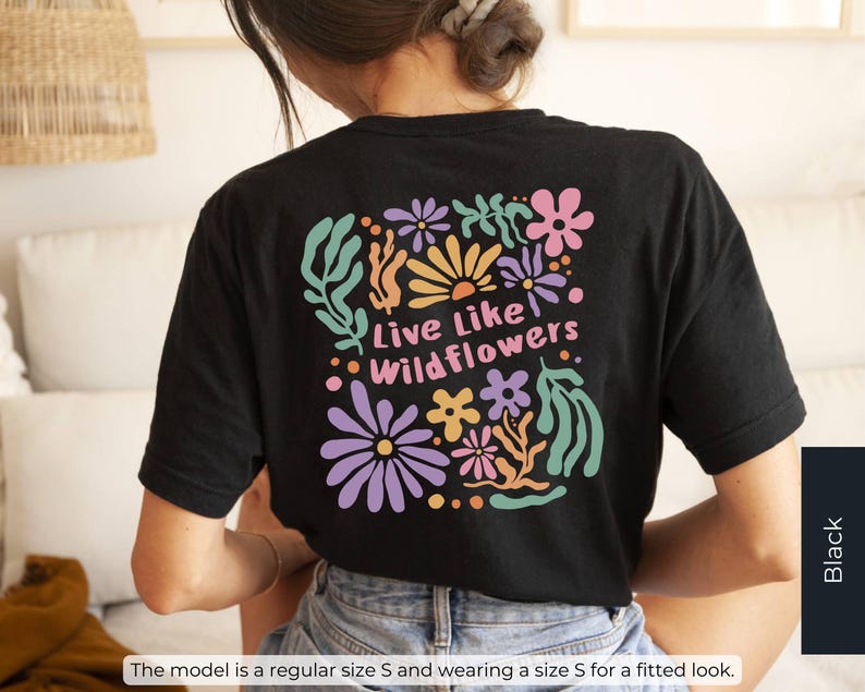 K&ouml;nnte beinhalten: Schwarzes T-Shirt mit einem bunten Blumendesign auf dem R&uuml;cken, mit dem Text "Live Like Wildflowers". Das Design umfasst Blumen in den Farbt&ouml;nen Rosa, Lila, Orange und Gr&uuml;n. Das Model tr&auml;gt Gr&ouml;&szlig;e S.