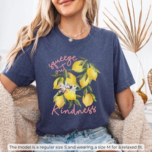 K&ouml;nnte beinhalten: Ein marineblaues T-Shirt mit einem Motiv aus Zitronen und Blumen. Der Text "squeeze out kindness" ist in Pink auf dem Shirt gedruckt.