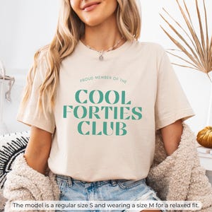 May include: A beige t-shirt with the text "PROUD MEMBER OF THE COOL FORTIES CLUB" in green lettering.