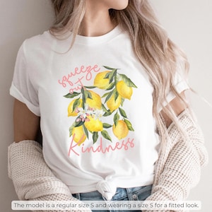 K&ouml;nnte beinhalten: Wei&szlig;es T-Shirt mit einem Motiv aus Zitronen und Bl&auml;ttern. Der Text "Squeeze out Kindness" ist in Pink auf dem Shirt gedruckt.