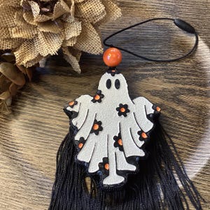 May include: A white ghost-shaped ornament featuring black and orange flower patterns, topped with a single orange bead and a black elastic loop. The bottom of the ghost is adorned with a long, dense black fringe made of thin, hanging threads.