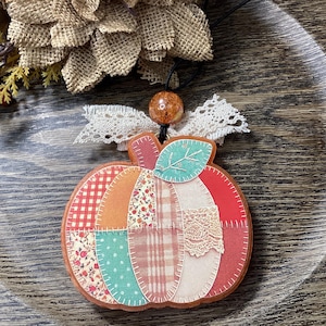 May include: A pumpkin-shaped ornament featuring a patchwork design with various patterns including red gingham, floral prints, and plaid. The ornament is accented with a teal leaf, a white lace bow, and a single brown bead on a black string.
