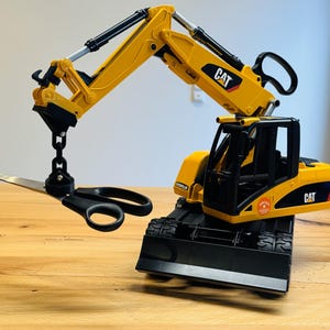 Könnte beinhalten: Ein gelber und schwarzer Spielzeugbagger mit dem CAT-Logo, der eine schwarze Schere hält. Der Bagger steht auf einer Holzoberfläche. Der Arm des Baggers ist ausgefahren, und die Schere hängt am Eimer.