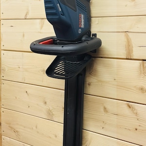 Könnte beinhalten: Eine blaue und schwarze elektrische Heckenschere von Bosch, montiert an einer Holzwand. Die Heckenschere hat ein rotes Sicherheitsmerkmal und einen schwarzen Griff. Die Klingen sind nicht sichtbar. Das Werkzeug ist zum Trimmen von Hecken und Büschen bestimmt.