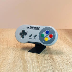 Könnte beinhalten: Ein grauer Super Nintendo Entertainment System-Controller mit einem schwarzen Ständer. Der Controller hat das klassische Tastenlayout mit A, B, X, Y, Start und Select-Tasten. Der Controller befindet sich auf einer Holzoberfläche.