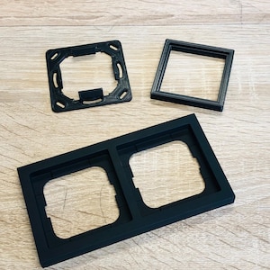 Könnte beinhalten: Drei schwarze Kunststoff-Steckdosenabdeckungen. Zwei der Abdeckungen sind rechteckig und haben zwei Öffnungen für Steckdosen. Die dritte Abdeckung ist quadratisch und hat eine Öffnung für eine Steckdose.