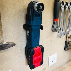 Könnte beinhalten: Ein kabelloses oszillierendes Multitool mit einem schwarzen und tealfarbenen Gehäuse und einem roten Akkupack. Das Werkzeug ist auf einer Holzoberfläche montiert, im Hintergrund sind weitere Werkzeuge zu sehen.