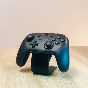 Könnte beinhalten: Ein schwarzer Nintendo Switch Pro Controller mit einem schwarzen Ständer. Der Controller hat ein schwarz-weißes Design mit den Tasten Kreuz, Kreis, X und Dreieck.