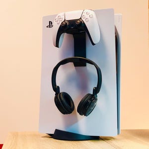 Könnte beinhalten: Eine weiße PlayStation 5 Konsole mit einem schwarzen Controller und schwarzen Kopfhörern, die an einem Ständer hängen. Die Konsole steht auf einer Holzoberfläche.
