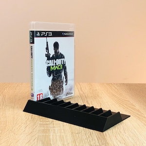 Könnte beinhalten: Ein schwarzer Plastik-Videospiel-Gehäusehalter mit einem PlayStation 3-Spielgehäuse oben drauf. Das Spielgehäuse ist für Call of Duty: Modern Warfare 3. Das Spielgehäuse ist schwarz und grün mit einem Soldaten auf der Vorderseite.