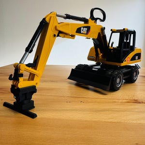 Könnte beinhalten: Ein gelb-schwarzes CAT-Bagger-Spielzeug mit schwarzer Schaufel und Rädern. Das Spielzeug hat eine Kabine mit schwarzem Griff und das CAT-Logo ist auf dem Arm und der Seite sichtbar.