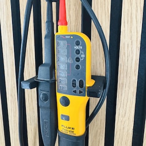 Könnte beinhalten: Ein gelb-schwarzer Fluke T110 VDE Spannungstester mit roter Spitze und schwarzen Kabeln. Das Gerät zeigt Spannungspegel an und verfügt über Tasten für AC- und DC-Spannungstests. Das Gerät hängt vor einem hölzernen Lattenhintergrund.