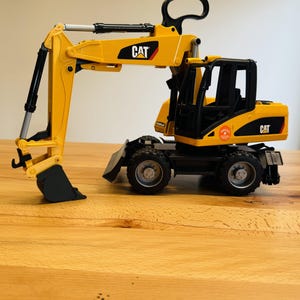 Könnte beinhalten: Ein gelb-schwarzes CAT-Bagger-Spielzeug mit schwarzem Griff und schwarzem Eimer. Das Spielzeug steht auf einer Holzoberfläche. Das CAT-Logo ist auf der Seite des Baggers zu sehen.