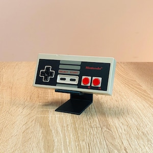 Könnte beinhalten: Ein klassischer Nintendo Entertainment System-Controller in Beige und Schwarz mit roten Knöpfen, auf einem schwarzen Ständer präsentiert. Der Controller trägt die Aufschriften "SELECT START" und "Nintendo".