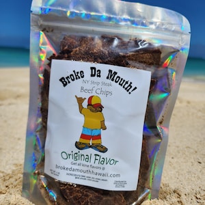 May include: A bag of Broke Da Mouth! NY Strip Steak Beef Chips in the Original Flavor. The bag is on a sandy beach with a blue sky and ocean in the background. The bag has a cartoon character with a surfboard and the text "Get all kine flavors @ brokedamouthhawaii.com".