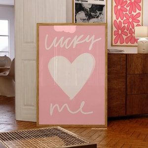 May include: Pink and white art print with a heart and the words "lucky me" in white script. The print is framed in a light wood frame and is hanging on a wall.