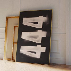 May include: A black framed art print featuring the number "444" in a white, distressed font. The artwork leans against a white wall, next to a framed canvas with a gold frame. The floor is made of light-colored wood.