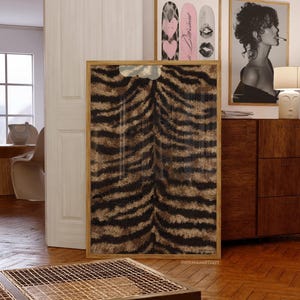 May include: A framed artwork featuring a tiger stripe pattern in shades of brown and black. The artwork is displayed in a room with other framed art, a wooden cabinet, and a table with a metal grid top.