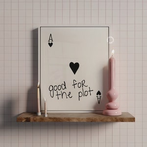 May include: A white framed print with the text "good for the plot" and an ace of spades playing card graphic. Two small candles and a pink candle are on a wooden shelf below the print.