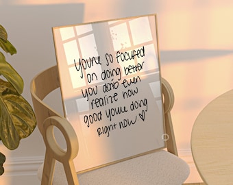 May include: A framed inspirational quote on a white background with black text that reads "You're so focused on doing better you don't even realize how good you're doing right now. ♡". The frame is sitting on a white chair with a wooden frame.