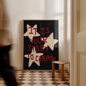 Könnte beinhalten: Ein gerahmter Kunstdruck mit schwarzem Hintergrund und dem Text "IT IS YOUR STORY TO CREATE" in Rot und Weiß. Das Kunstwerk zeigt drei weiße Sternformen. Ein kleiner Holzschemel steht in der Nähe auf einem karierten Boden.