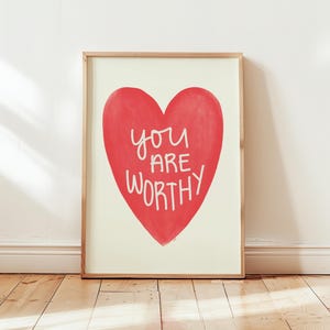 May include: A framed print with a red heart and the text "you are worthy" in white lettering.