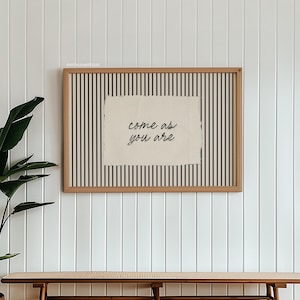 May include: A framed artwork with the text "come as you are" on a textured background, set against a black and white striped pattern. The artwork is displayed on a white paneled wall above a wooden bench.