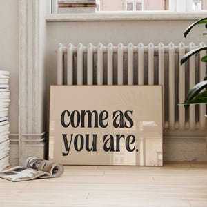 May include: A framed print with the words "come as you are" in bold black letters on a beige background. The print is leaning against a wall in a room with a window, radiator, and a potted plant. A stack of books and magazines are also visible.