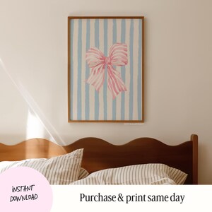 May include: A printable digital download of a pink bow on a blue and white striped background. The image is framed and hanging on a wall above a wooden bed with striped bedding.