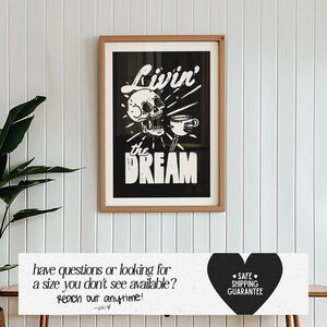 May include: A framed black and white art print with the words "Livin' the DREAM" and a skull graphic. The print is in a light brown frame and hangs on a white wall. The art print also includes a coffee cup graphic.