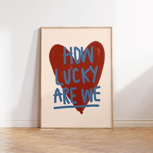 May include: A framed art print featuring a large red heart with the blue text "HOW LUCKY ARE WE". The artwork has a minimalist style with a cream-colored background and a wooden frame.