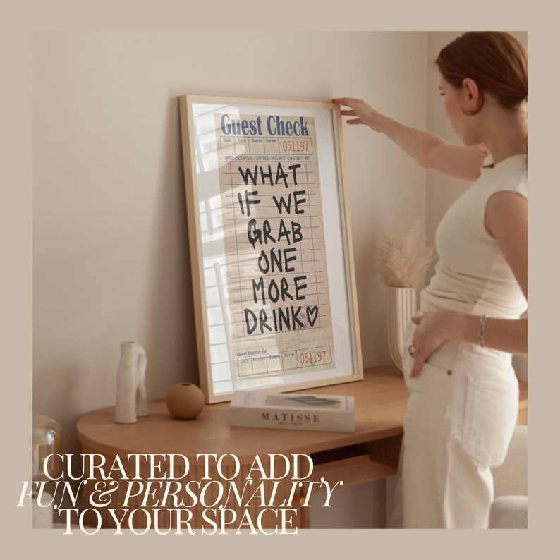May include: A framed print with a white background and black text that reads "Guest Check" and "What if we grab one more drink". The print is hanging on a wall above a wooden table with a book and a small vase.