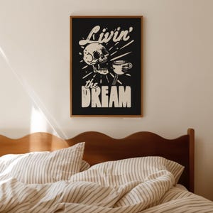 May include: A framed black and white art print featuring a skull holding a coffee cup. The text "Livin' the Dream" is displayed above and below the skull. The artwork is in a wooden frame and hangs above a bed.