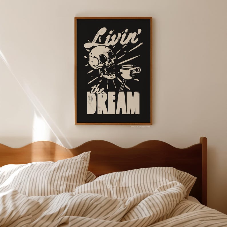 May include: A framed black and white art print featuring a skull holding a coffee cup. The text "Livin' the Dream" is displayed above and below the skull. The artwork is in a wooden frame and hangs above a bed.